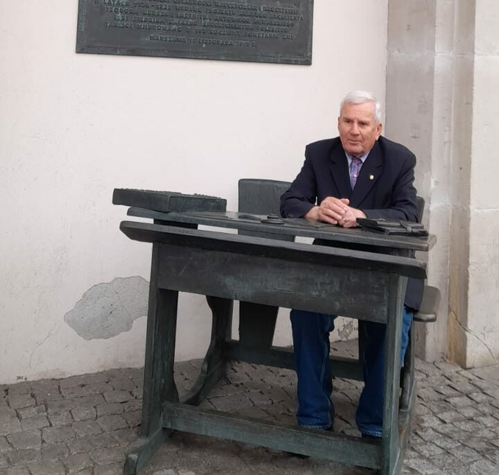 Jerzy Szmytka w ławce szkolnej – replice ławki z 1905 roku, przed Domem Nauczycielskim na warszawskim Powiślu. Na ścianie budynku widoczna pamiątkowa tablica, wmurowana i odsłonięta w 2010 roku (Fot. archiwum prywatne).